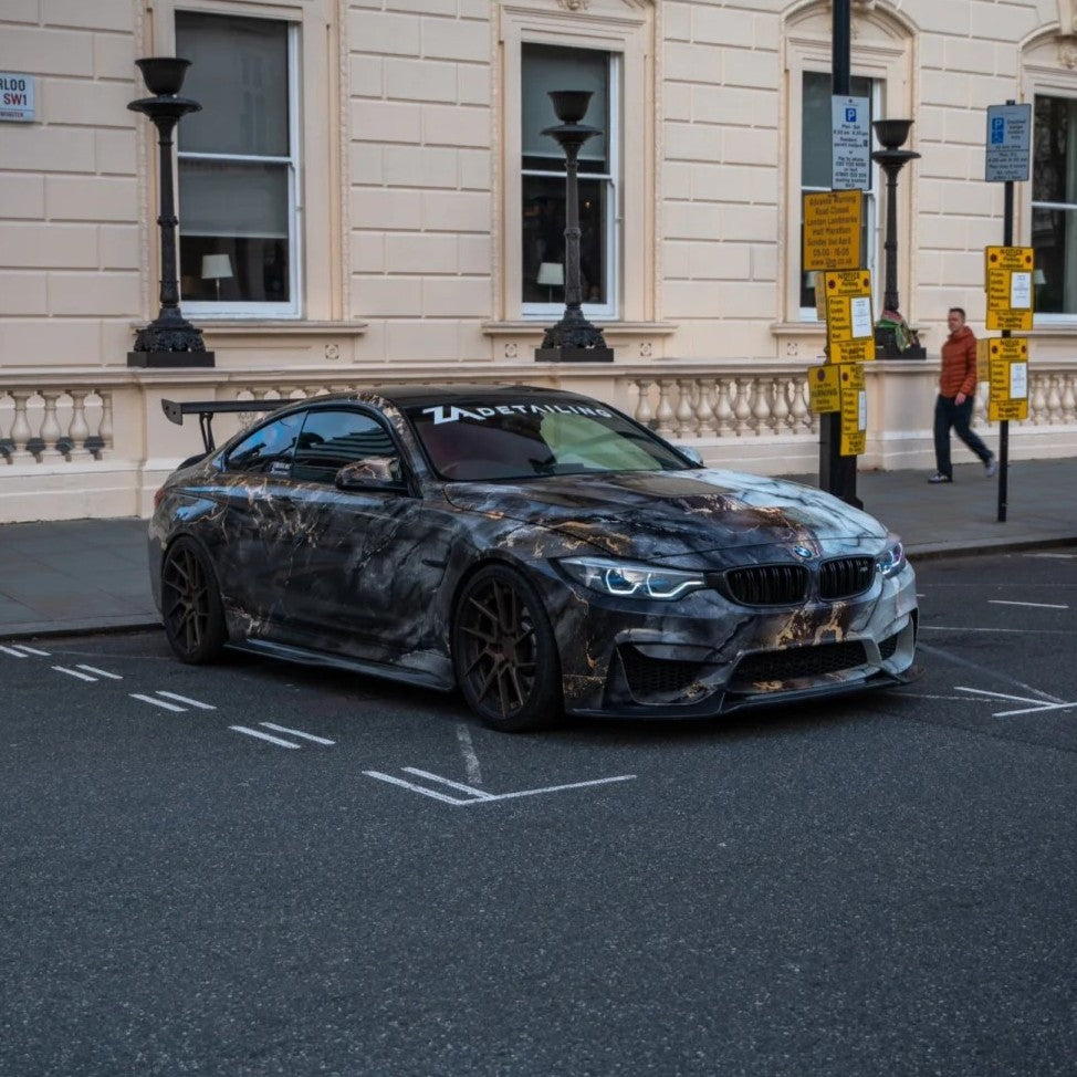 A customized BMW M2 coupe with a dark gray and black marble-patterned wrap, parked on a city street in front of a cream-colored building.